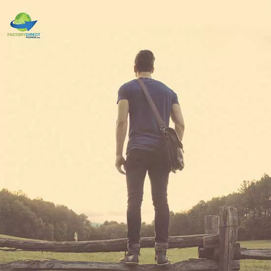 Man standing on a wooden fence with a custom messenger bag over his shoulder looking across an open field.