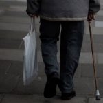 Elderly person with a cane walking on a sidewalk carrying a reusable bag